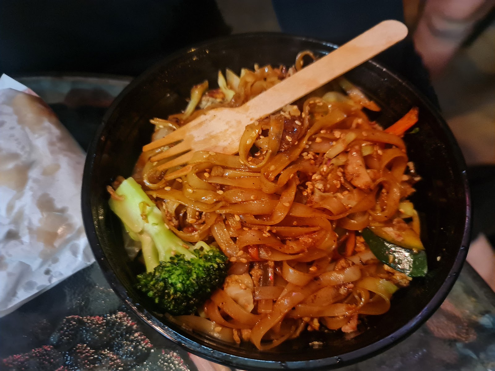 A bowl of wide rice noodles with sesame, broccoli and a wooden fork